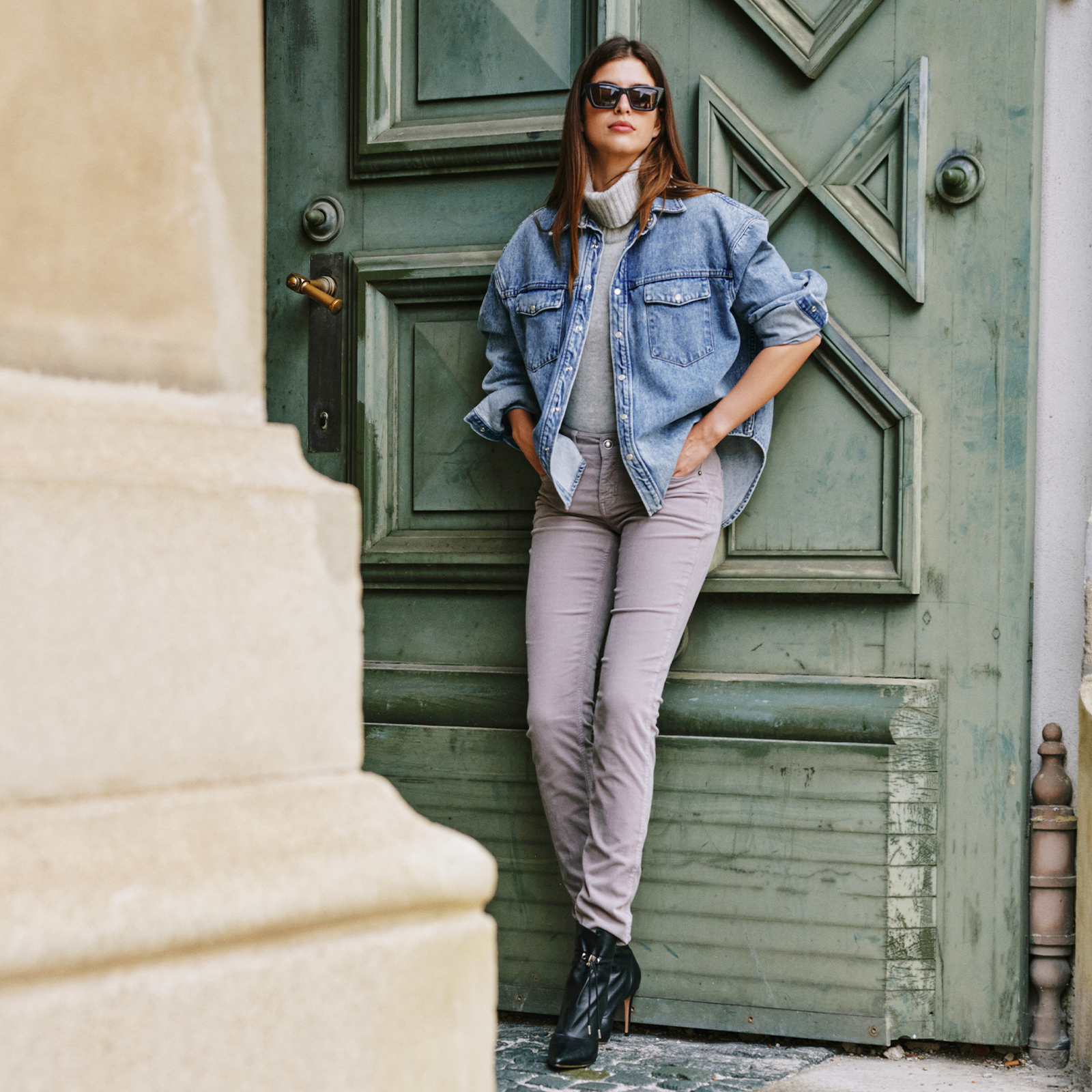 Woman wearing a denim jacket and slim-fit cozy denim, leaning against a green door outdoors.