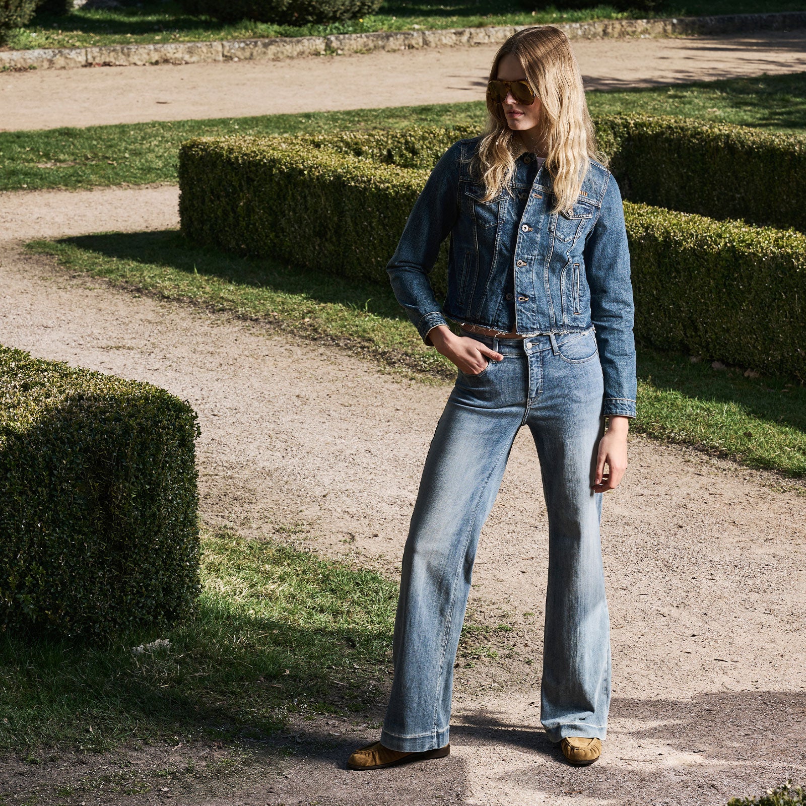 Woman wearing light blue flared jeans and a matching denim jacket, styled outdoors in a garden setting.