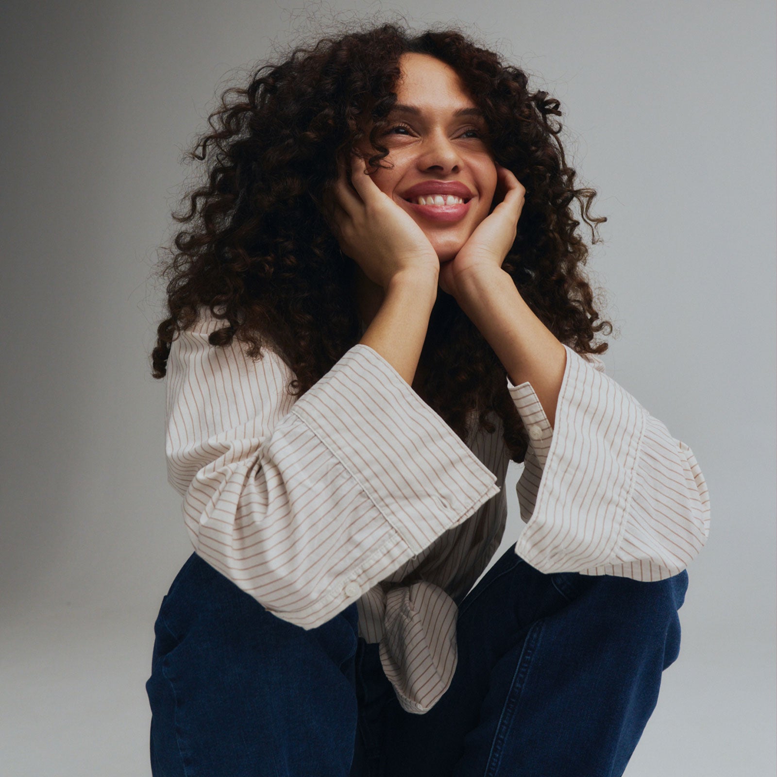 Smiling woman with natural curly hair wearing dark blue jeans and a light striped blouse seated in a relaxed pose.
