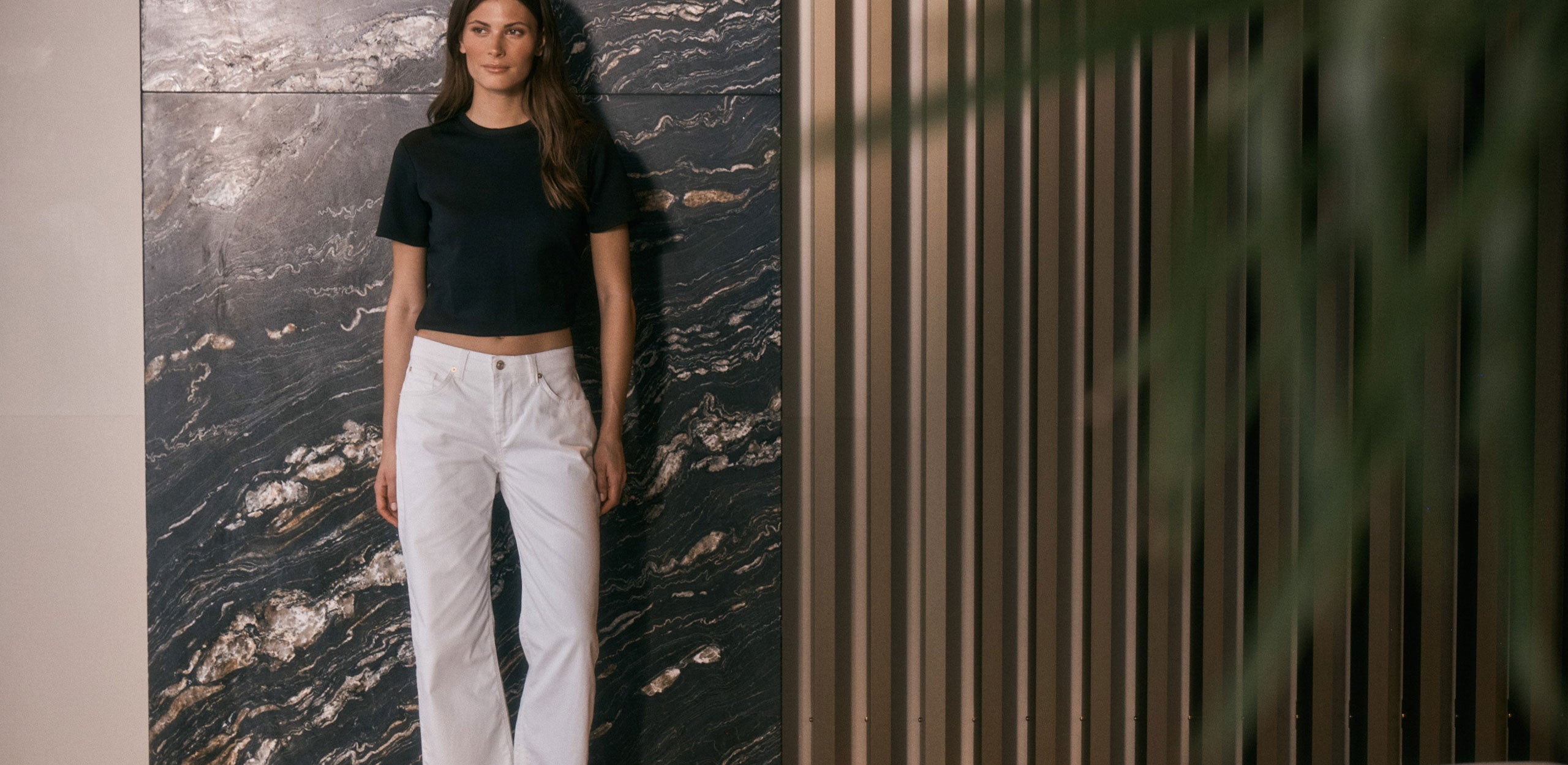 A woman wearing white straight-leg jeans and a black cropped top stands against a dark marble wall, creating a clean and modern contrast.