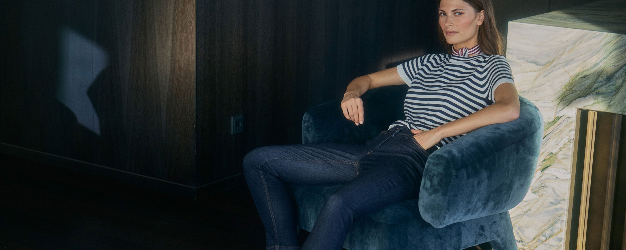 Woman wearing dark dream jeans and a striped top, sitting in a blue armchair in a softly lit interior.