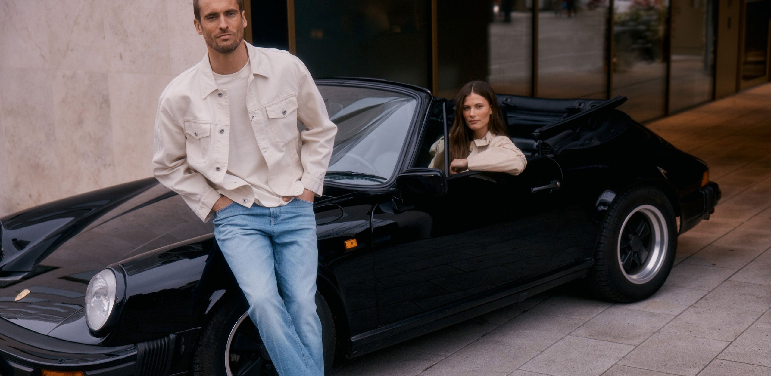 A man in a light jacket and blue jeans leaning against a black convertible, with a woman seated inside the car, parked in front of a modern building.