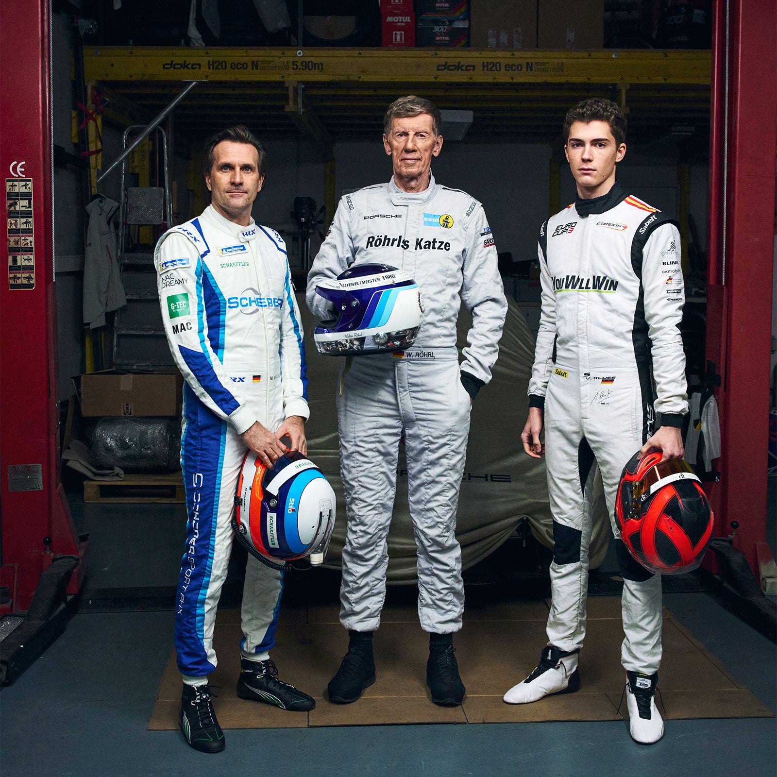 Three race drivers in white racing suits standing in a garage, each holding a helmet in front of a race car.