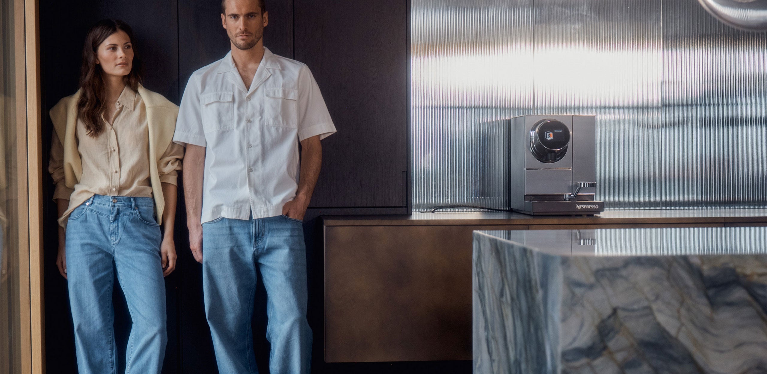 Two models wearing light blue denim jeans stand in a modern interior next to a coffee machine, styled in neutral spring outfits.