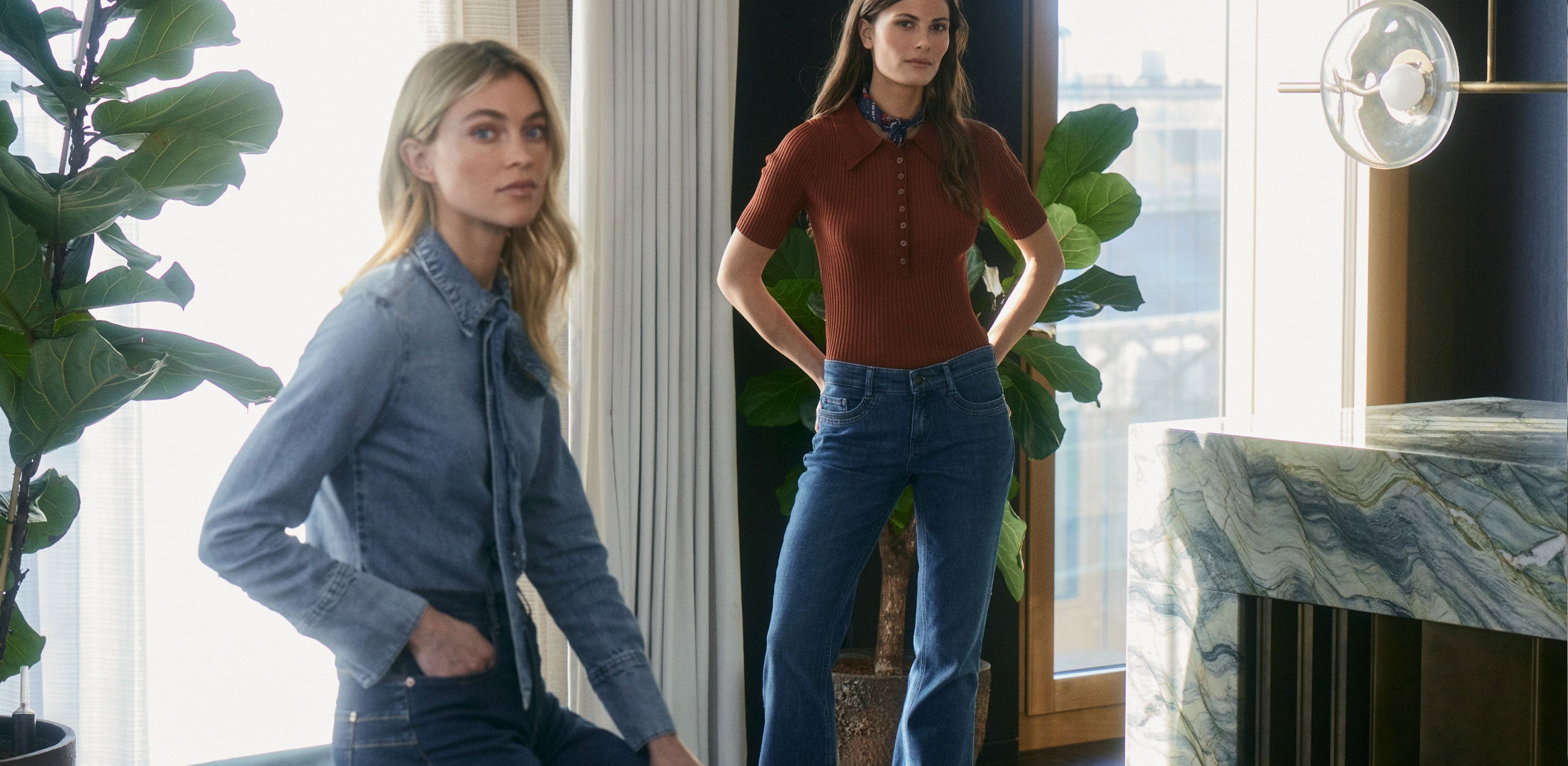 Two women modeling casual denim outfits indoors. On the left, a blonde woman wears a light blue chambray shirt with a tie-neck detail and dark jeans, posing with one hand in her pocket. On the right, a brunette woman stands in front of a window and a large green plant, wearing a fitted rust-colored short-sleeve knit top, blue jeans, and a neck scarf, with her hands on her hips. The setting is bright and modern with natural light.