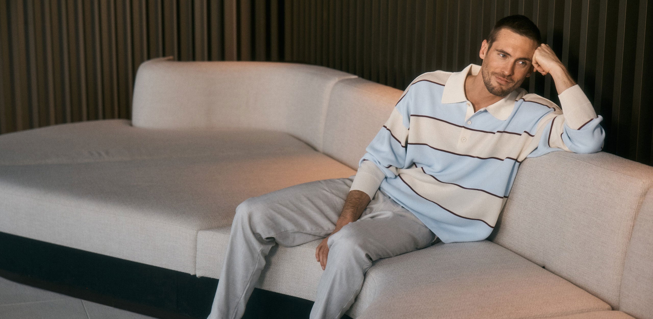 Man sitting on a beige sofa wearing a light blue striped knit polo and light trousers in a modern interior.