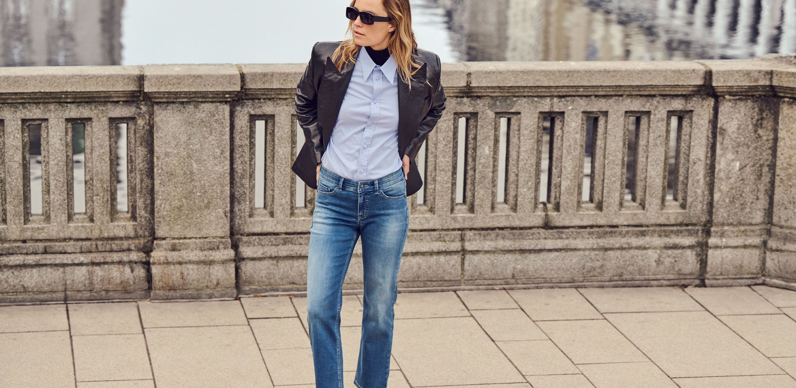 A person wearing a black blazer over a light blue button-up shirt, relaxed-fit blue jeans, white socks, and black loafers, standing by a stone railing near the water.