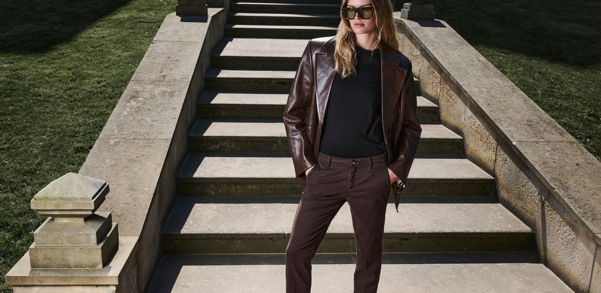 Woman wearing brown chino pants with a matching brown leather jacket, black top and sunglasses, standing on outdoor stone steps.