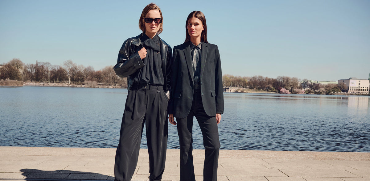 Two women wearing elegant all-black outfits with wide-leg and straight-leg pants, styled with a leather jacket and blazer, standing by a lake.