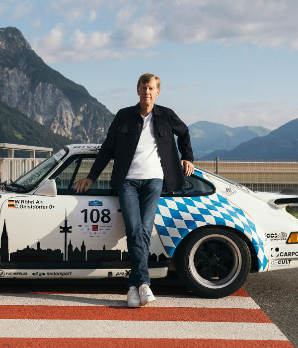 Man wearing a black jacket, white T-shirt, blue jeans and white sneakers, leaning against a classic racing car with blue and white detailing in front of a mountain landscape.
