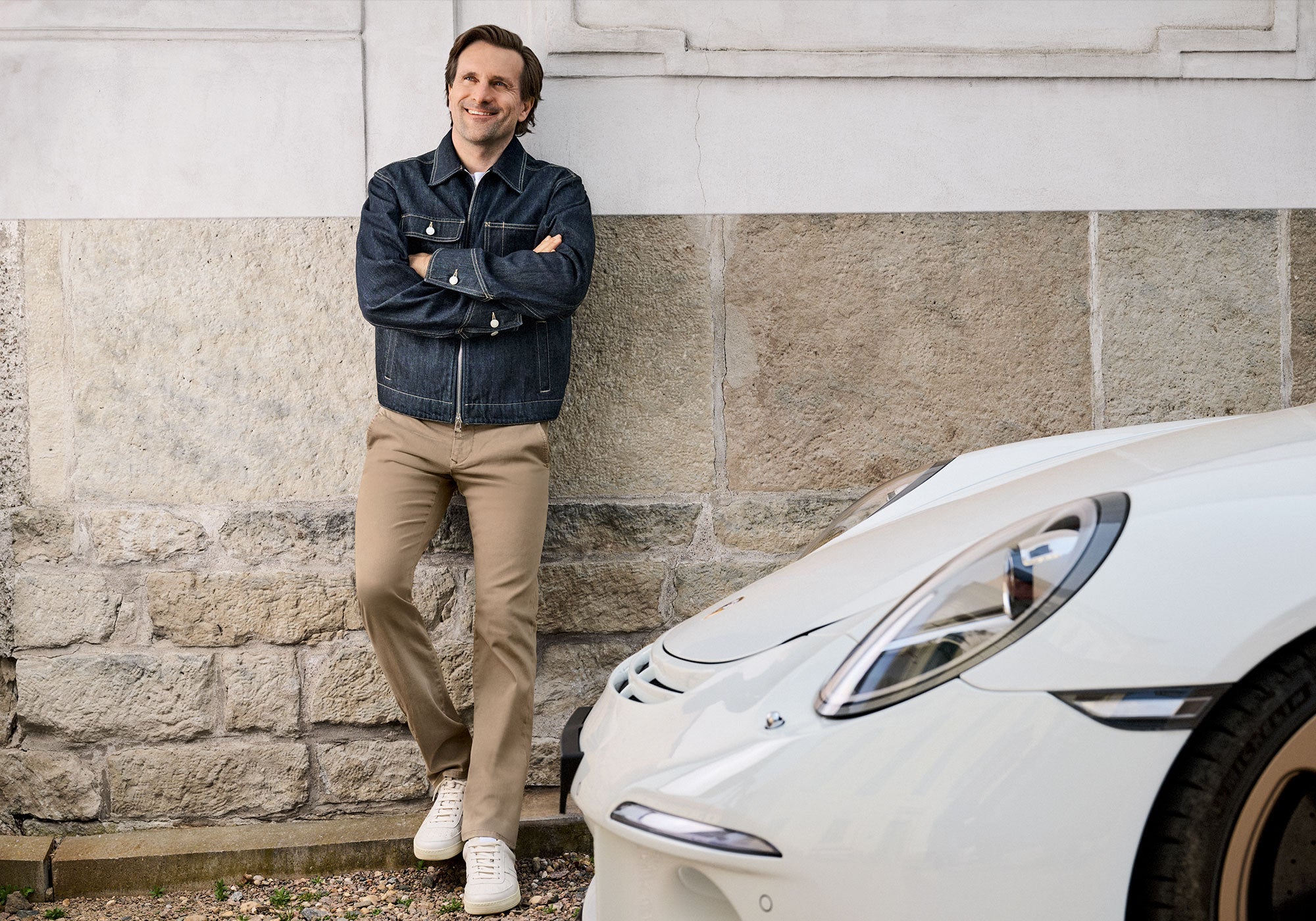 A man in a denim jacket and beige trousers leans casually against a wall beside a white car.