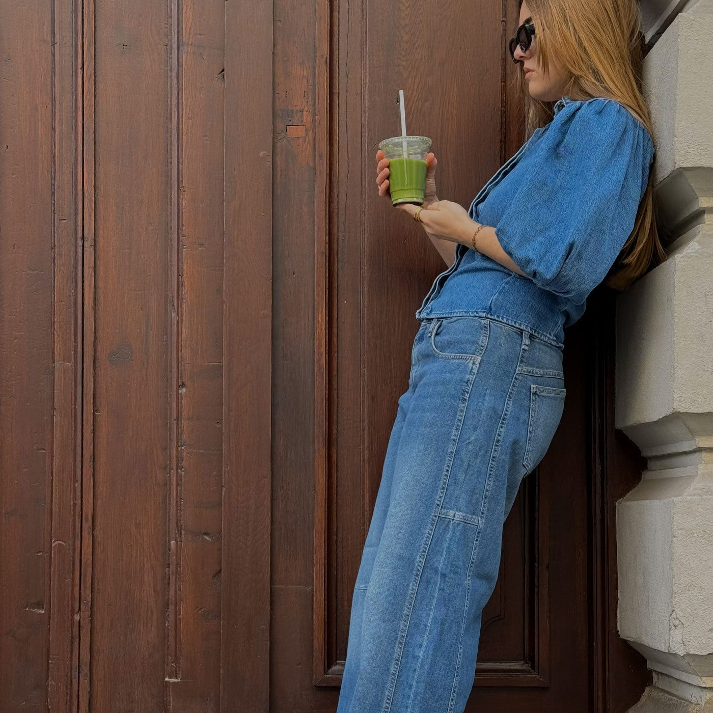 Woman in a blue denim outfit leaning against a wooden door while holding an iced matcha drink.