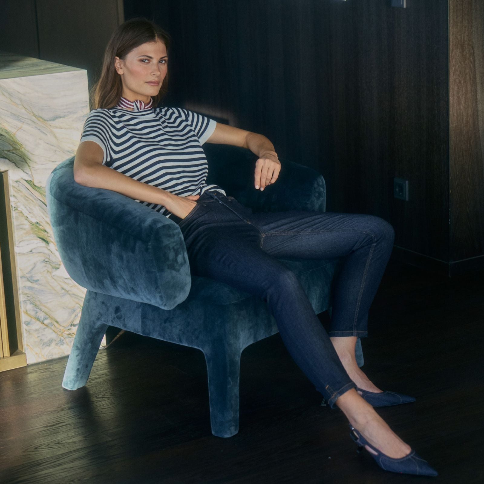 A woman wearing a navy and white striped short-sleeve top and dark skinny jeans sits relaxed in a blue velvet armchair, paired with navy flats, in a modern interior with dark walls and a marble side table.