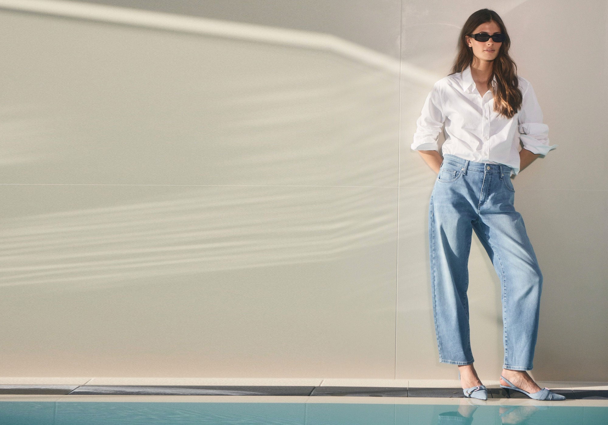 Woman in light blue denim jeans and white shirt standing by a pool in sunlight.