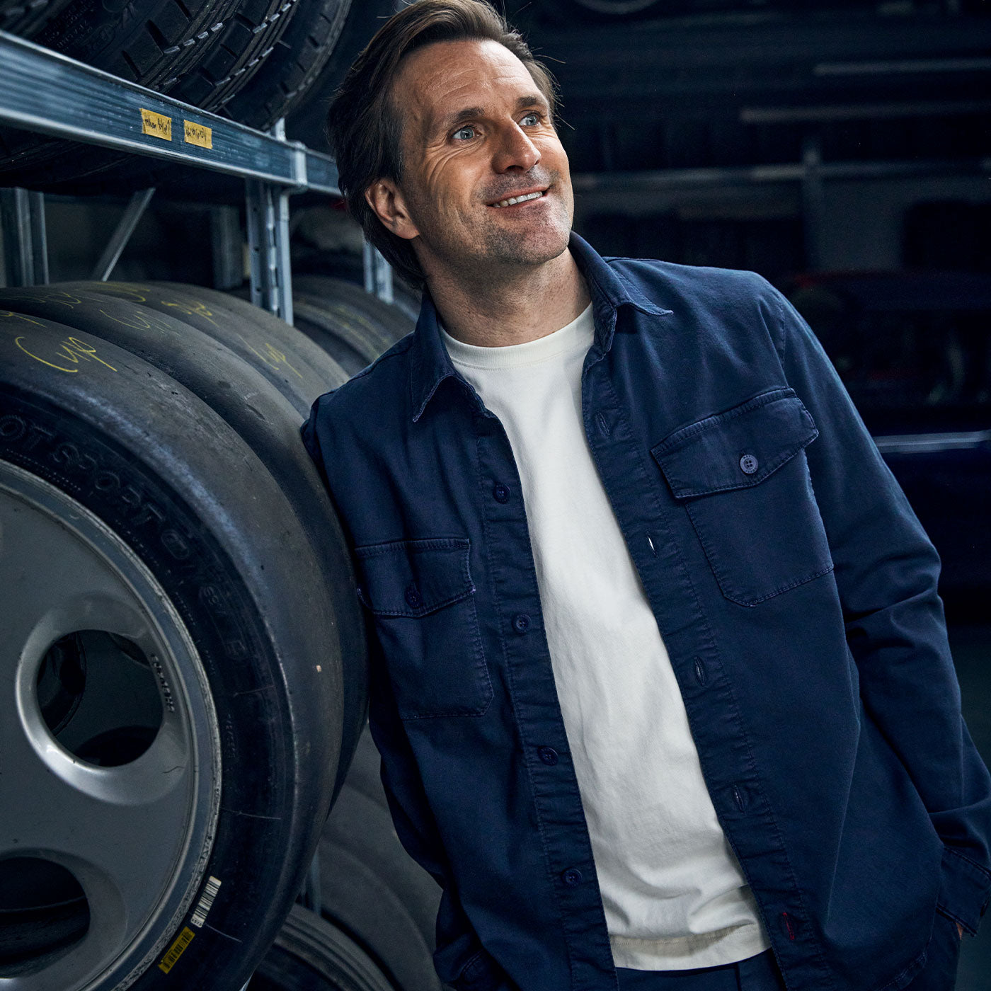 Man wearing a dark blue denim jacket and white T-shirt stands in a garage next to stacked racing tires, smiling slightly and looking off to the side.