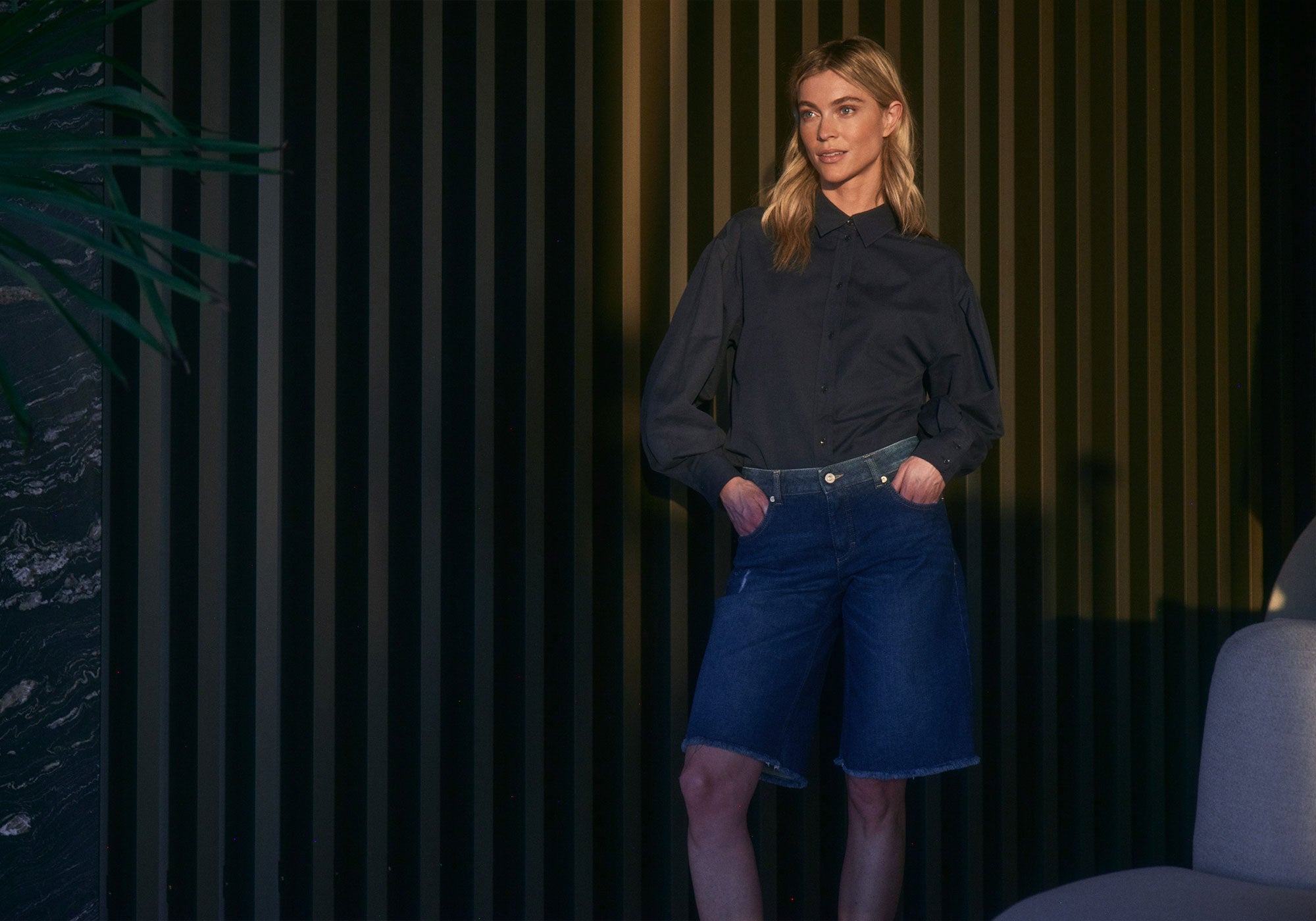A woman stands confidently in a modern indoor setting with dark vertical paneling. She wears a dark long-sleeve blouse tucked into knee-length denim Bermuda shorts. Her hands rest casually in her pockets, and she looks slightly to the side with a relaxed, self-assured expression. Soft lighting highlights her outfit, while a plant and part of a chair frame the scene, adding depth and a contemporary atmosphere.