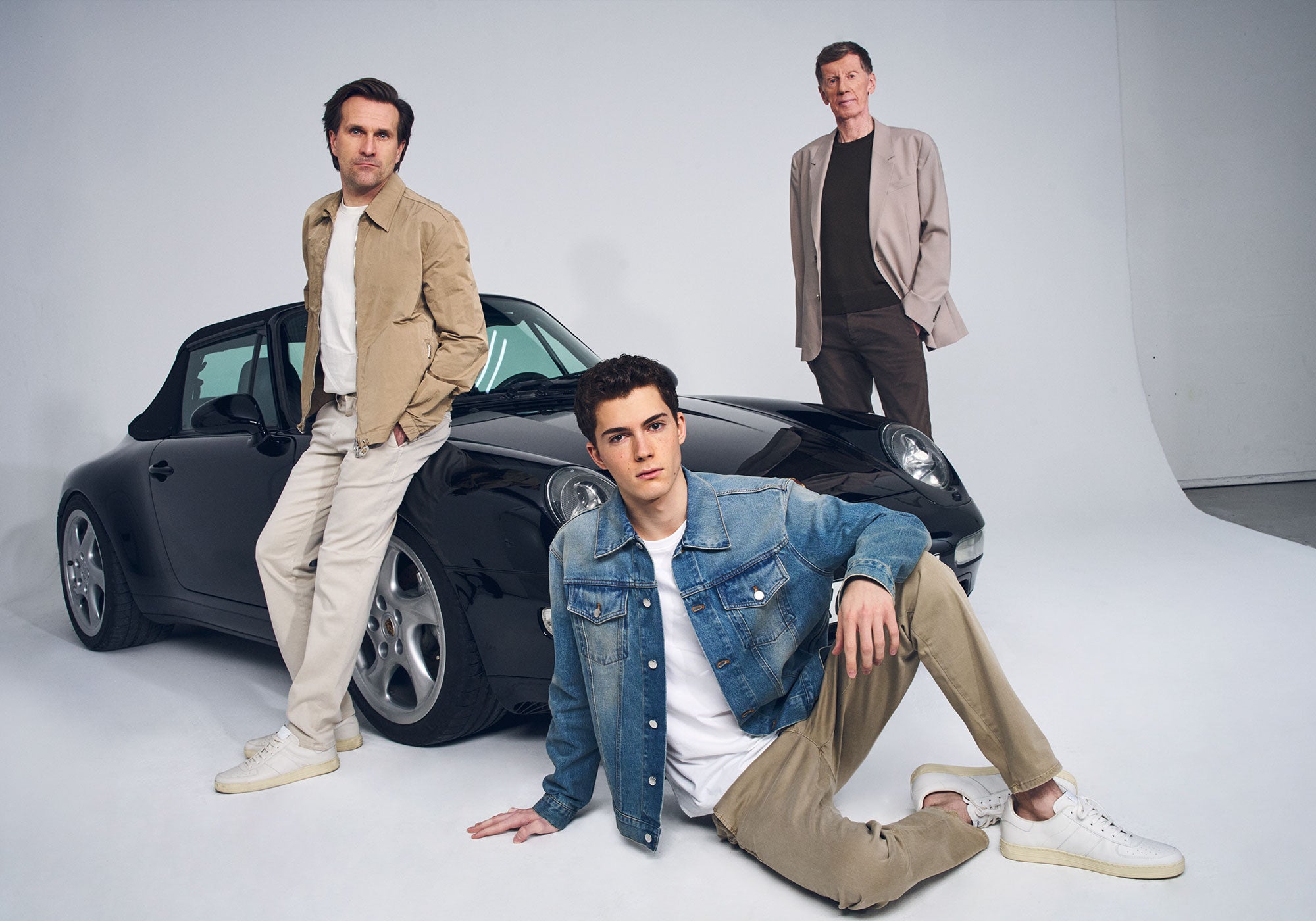 Three men from different generations pose around a black convertible sports car in a bright studio. One sits on the floor in a denim jacket, another leans against the car in a beige outfit, and the third stands behind the car wearing a blazer.