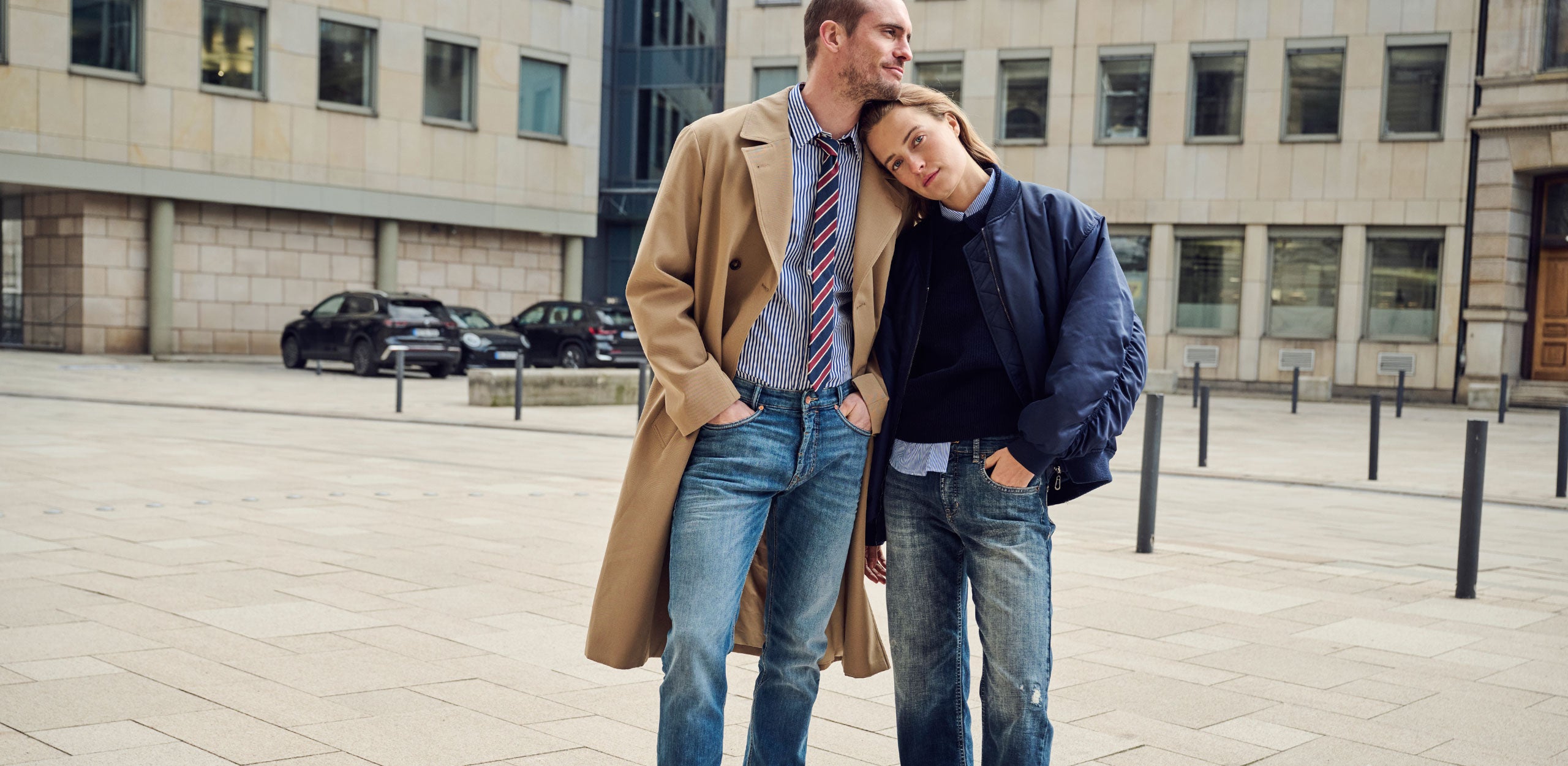 A casually dressed couple stands together in an open urban plaza. The woman rests her head on the man's shoulder as both look off into the distance. He wears a camel overcoat layered over a striped shirt, tie, and blue jeans, while she wears a navy bomber jacket, blue shirt, and relaxed-fit jeans. Modern office buildings and a few parked cars form the background.