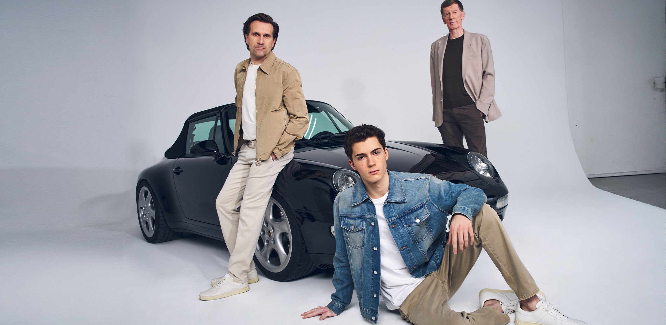 Three men wearing casual outfits with denim and neutral tones posing in front of a black convertible car.