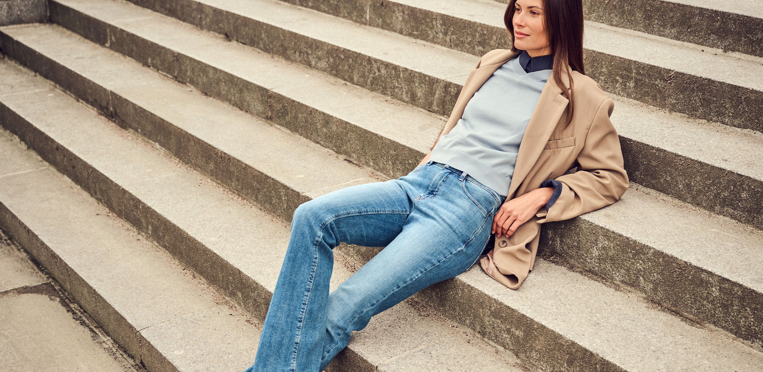 Woman wearing light blue jeans and a beige blazer, sitting on concrete steps.