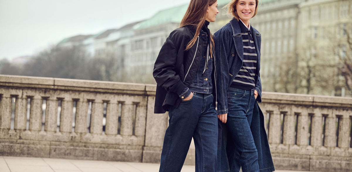 Two women wearing dark blue jeans and casual denim outfits with jackets and coats on a city street.