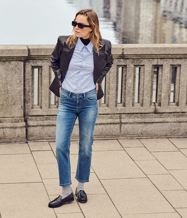 Woman wearing blue straight-leg jeans, a light blue shirt, black leather blazer, sunglasses and black loafers, posing by a stone railing.