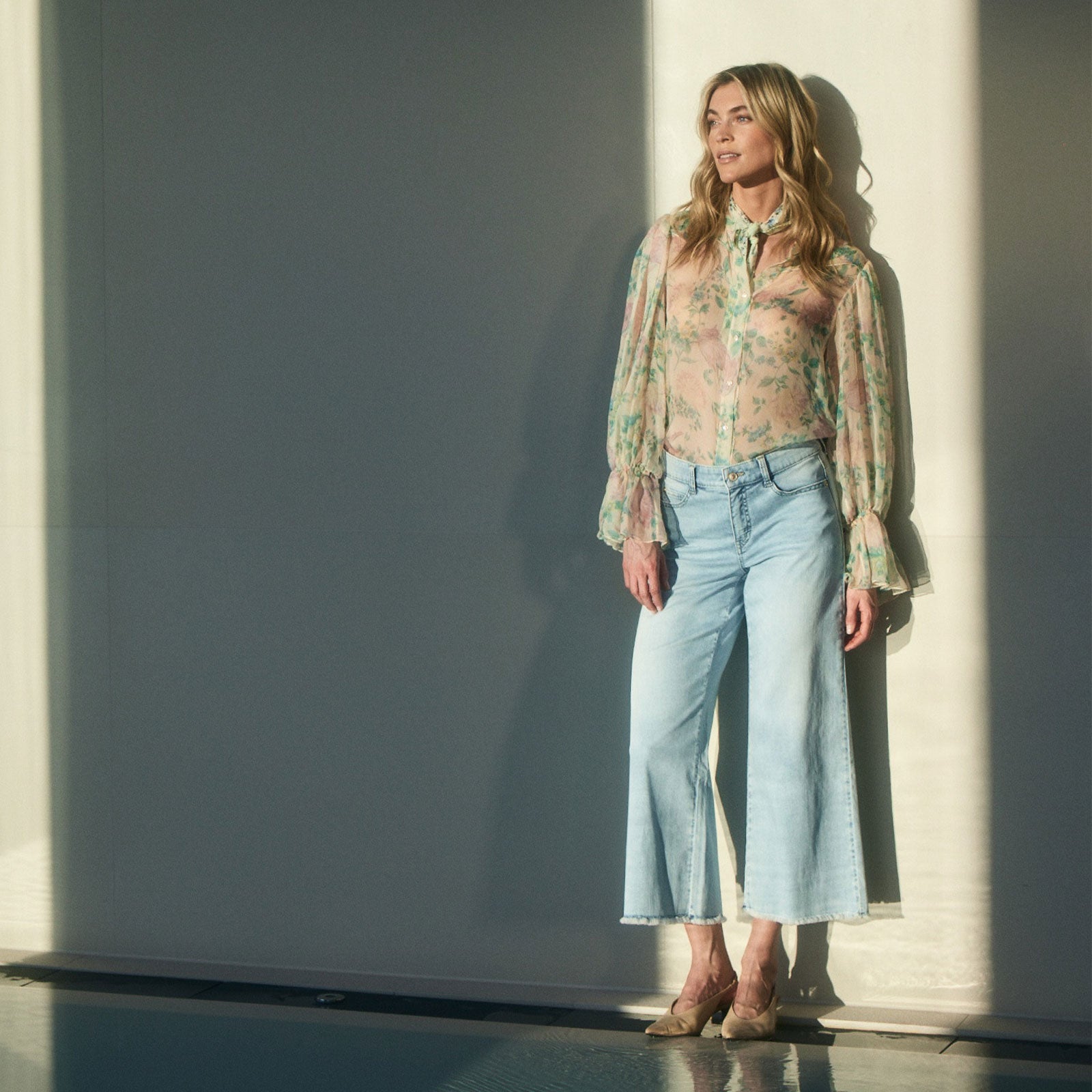 Woman standing in sunlight wearing a sheer floral blouse with long sleeves and light blue wide-leg cropped jeans, styled with beige heels, leaning against a wall with soft shadows creating a calm, elegant fashion scene.
