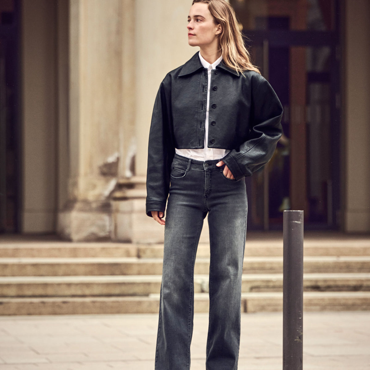 Woman wearing a cropped black leather-look jacket with grey Dream wide-leg jeans in an urban setting.
