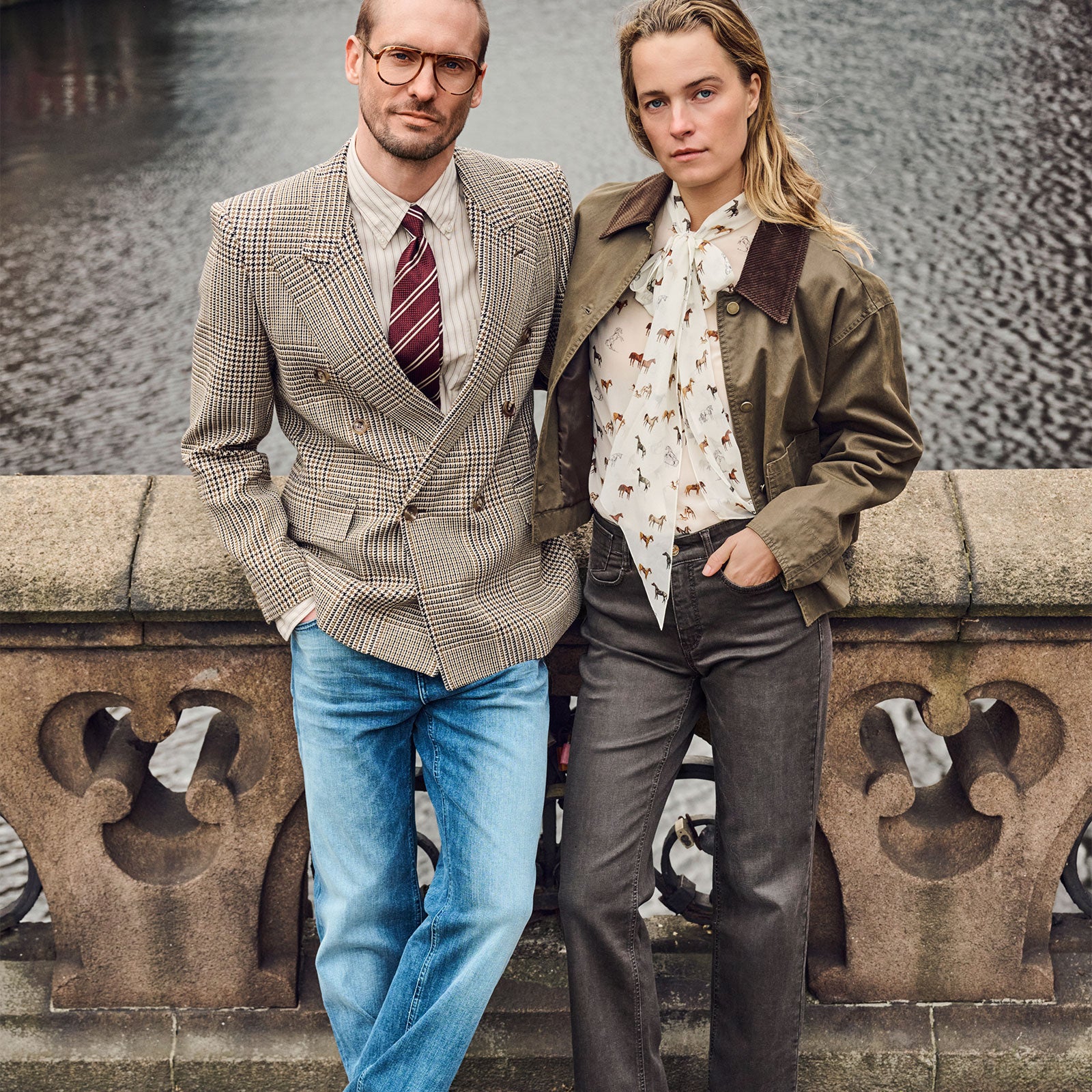 Man and woman wearing stylish casual outfits with jeans, posing together by a stone railing near the water.