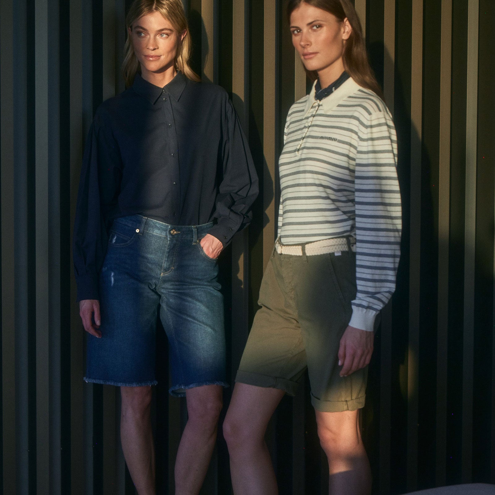 Two women wearing casual summer outfits—one in a dark blouse with denim shorts, the other in a striped top with olive shorts—posing in soft sunlight against a modern striped wall.