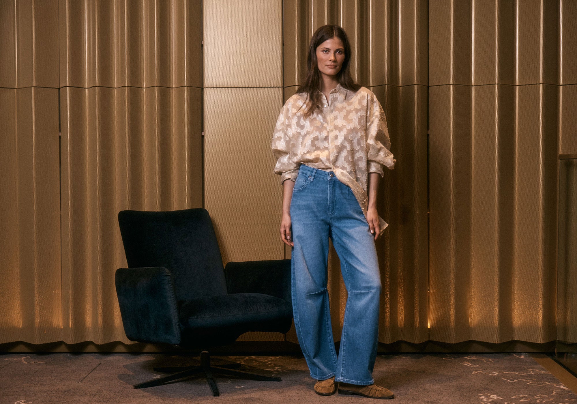 A woman in a beige blouse and blue wide-leg jeans stands beside a dark armchair against a golden textured wall.