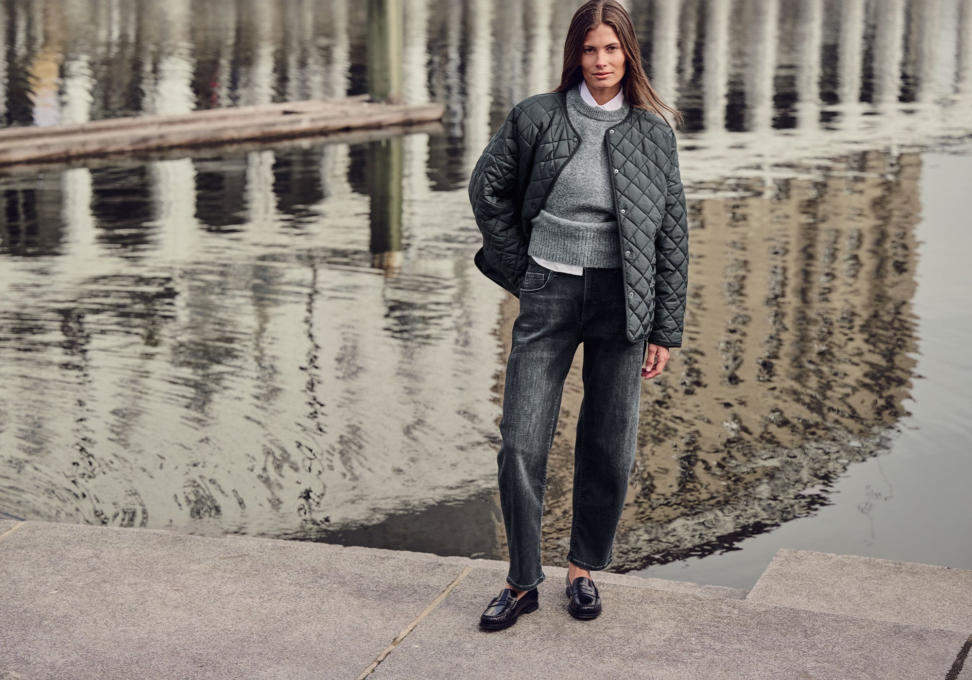 Women's model wearing a grey quilted jacket, knit sweater and dark barrel jeans by the waterfront.