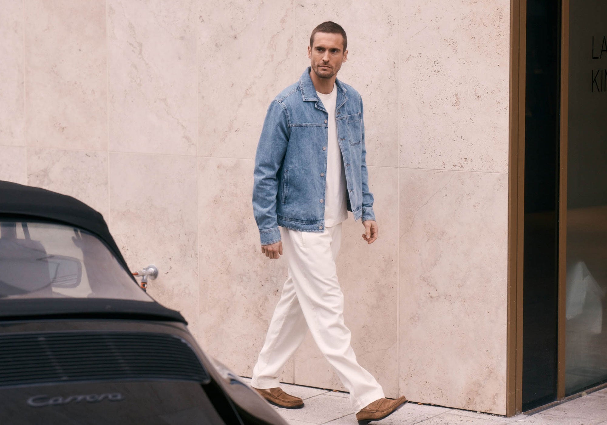 A man walking outdoors wearing light beige trousers, a white T-shirt, and a blue denim jacket, styled in a clean, modern spring look against a neutral stone wall.