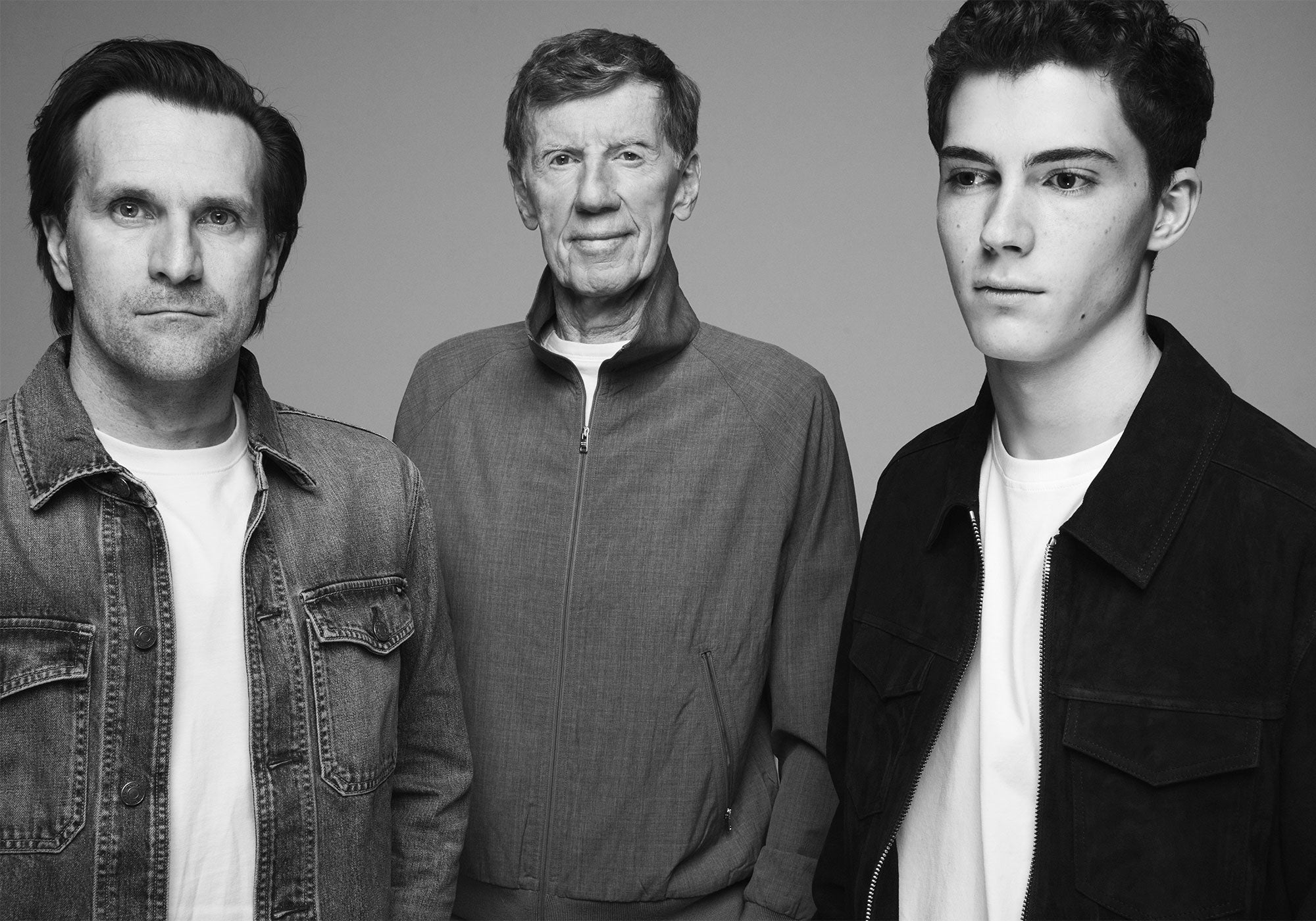 Black-and-white portrait of three men from different generations standing side by side against a plain background, wearing casual jackets and looking toward the camera.