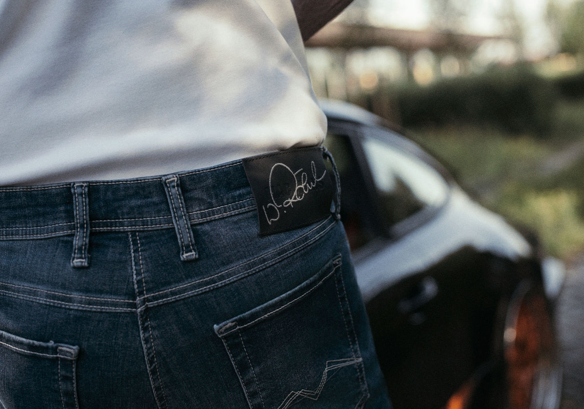 Close-up of MAC Jeans denim featuring a signature label, with a sports car in the background.