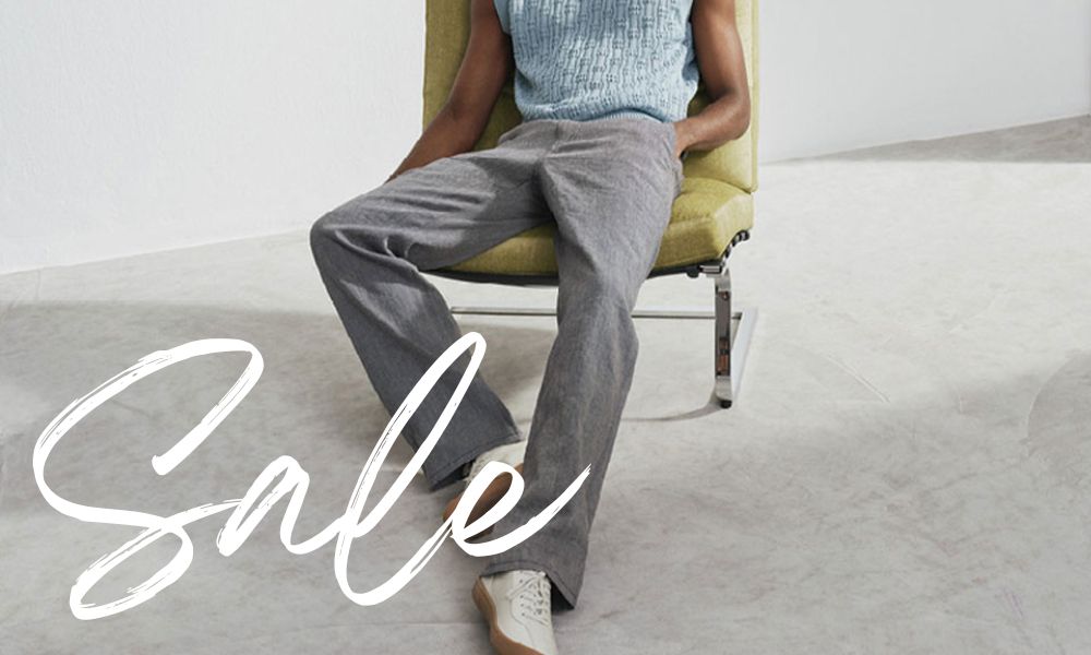 Men wearing light grey straight-leg trousers and white sneakers, seated in a modern upholstered chair in a minimalist interior with a light-toned floor and soft neutral colors.