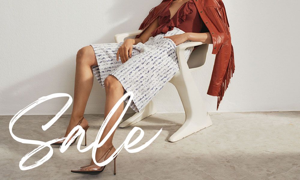 Woman seated in a modern white chair wearing a white patterned midi dress, a rust-colored fringed leather jacket, and transparent high heels in a minimalist interior with soft neutral tones.