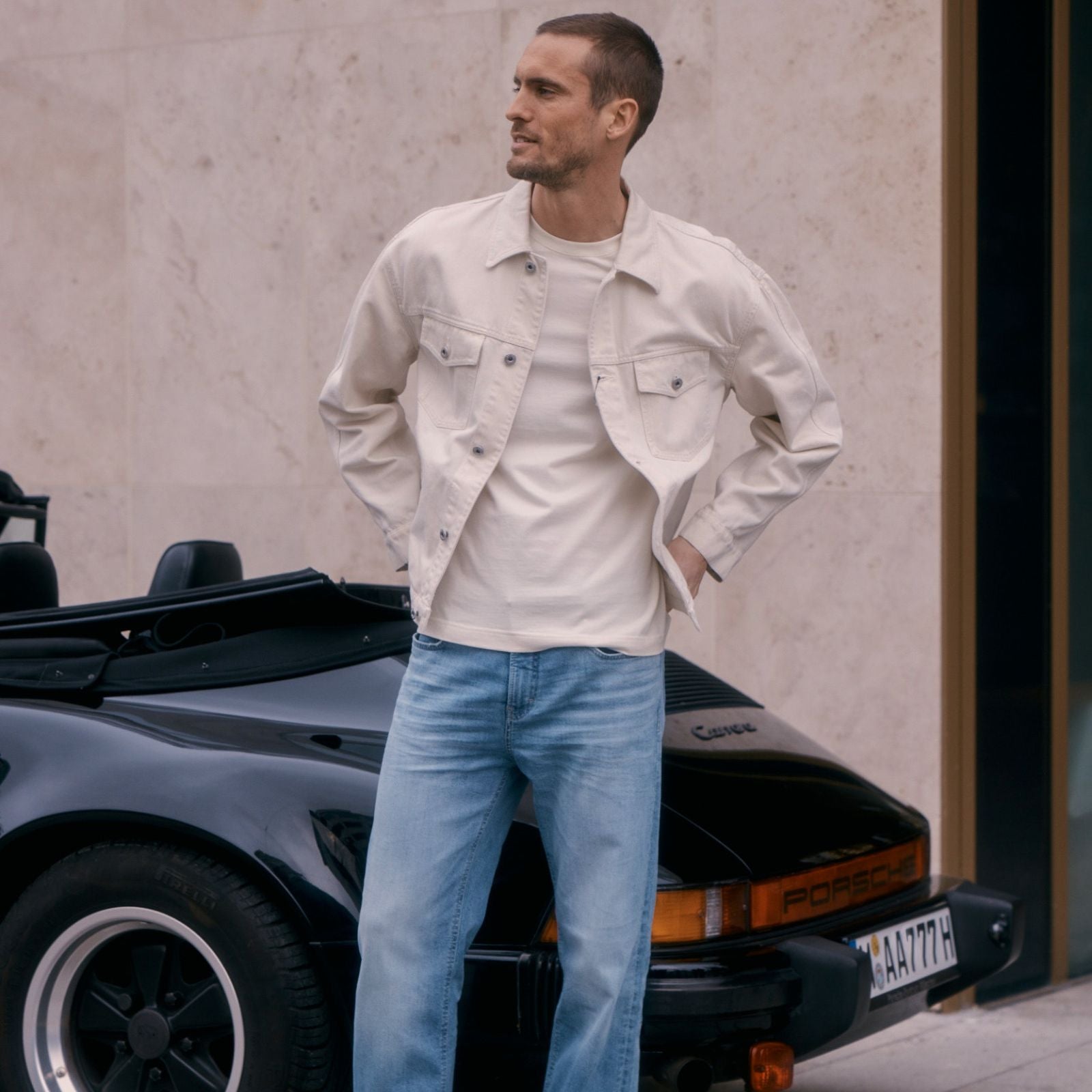 A man wearing a light beige denim jacket over a cream T-shirt and light blue jeans stands with his hands in his pockets beside a black convertible sports car parked in front of a modern beige stone wall.