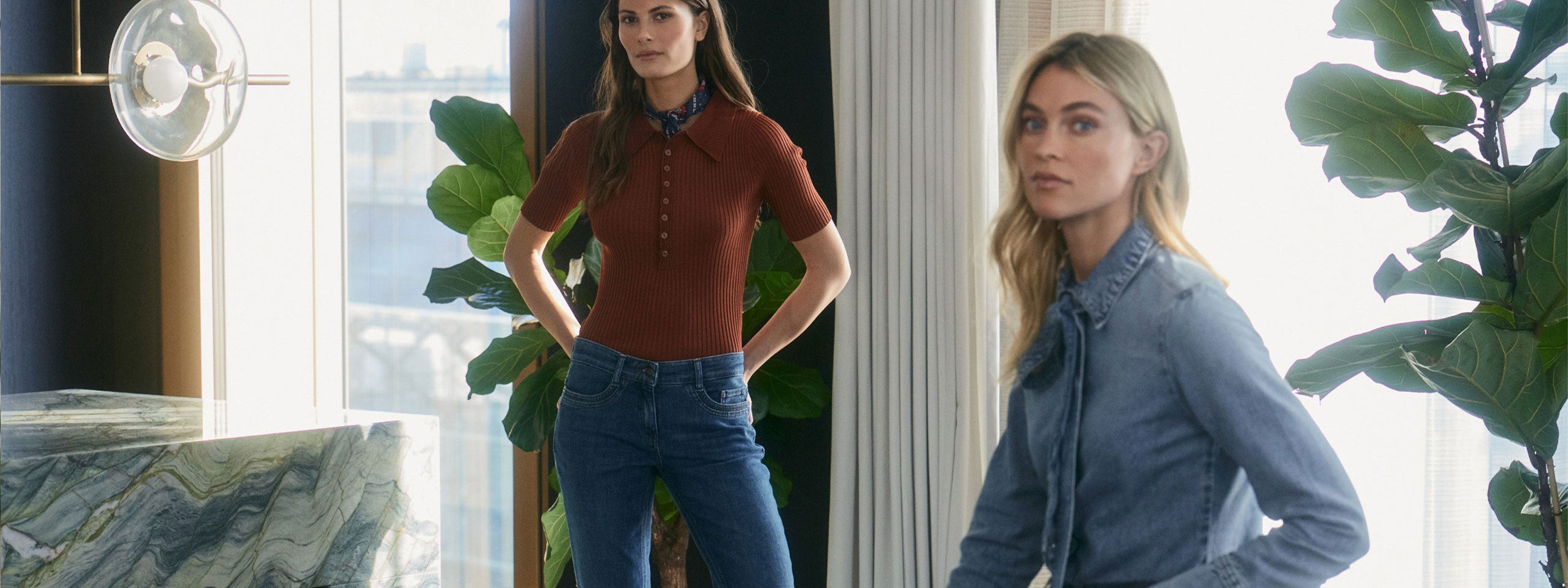 Two women wearing modern denim looks in a bright interior with large plants and natural light.