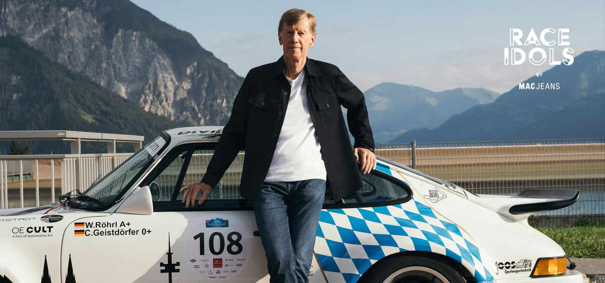 Walter Röhrl wearing MAC Jeans standing beside a classic race car in a mountain setting – Race Idols by MAC Jeans.