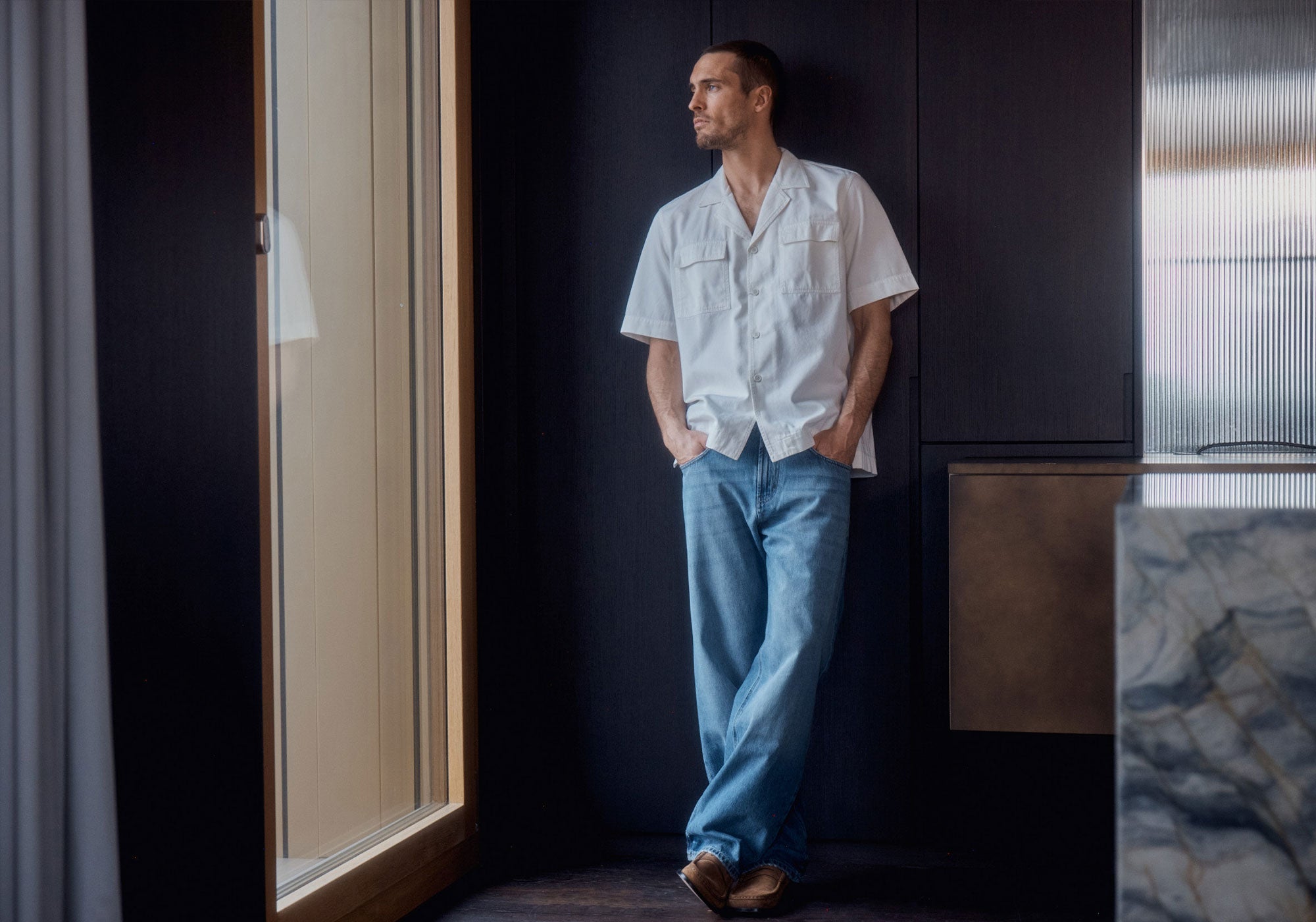 A man in a light short-sleeve shirt and blue jeans leans casually against a wall in a modern interior.