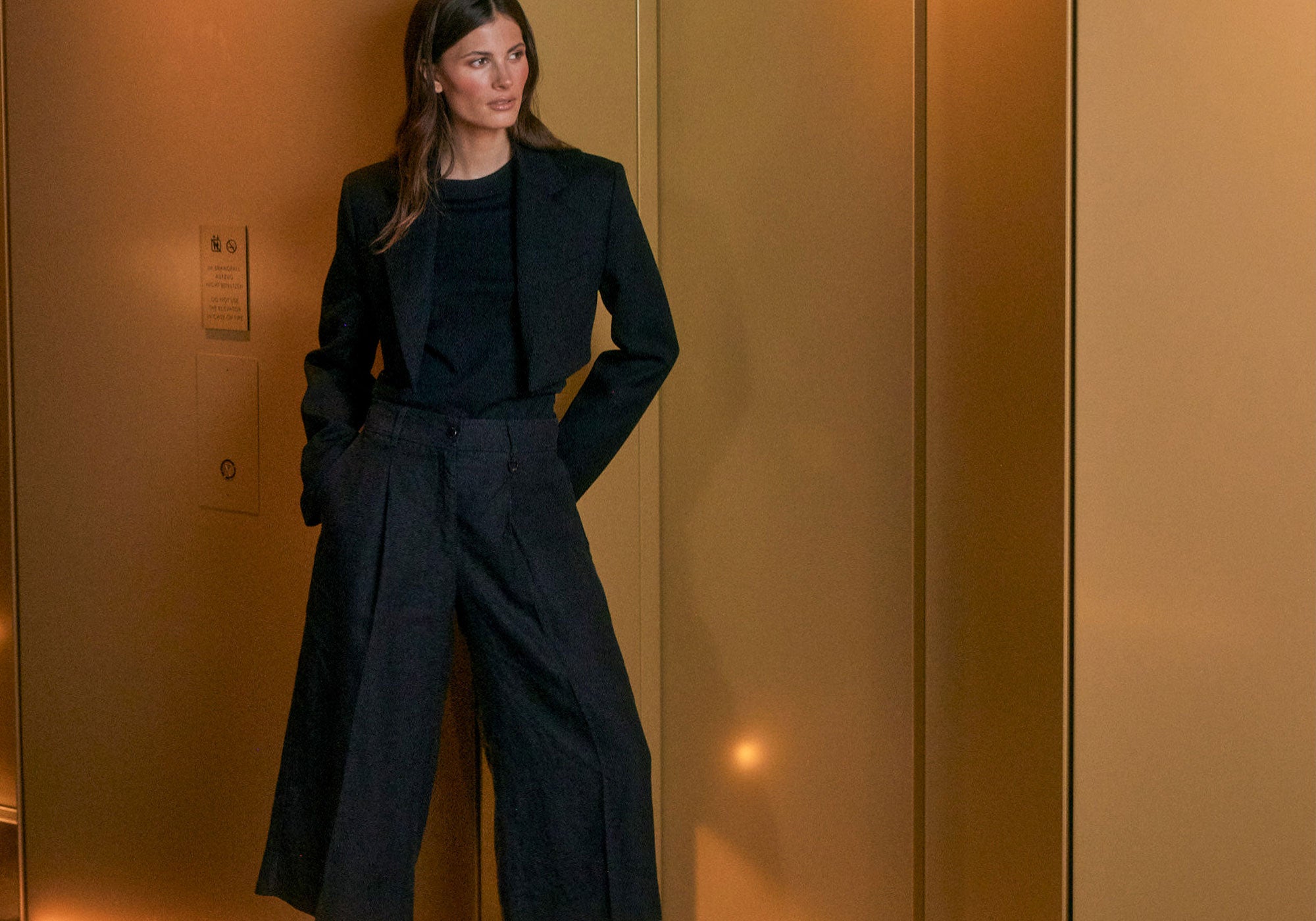 Woman wearing a dark tailored outfit with a cropped blazer and wide-leg trousers, standing in a warm-toned, modern interior.