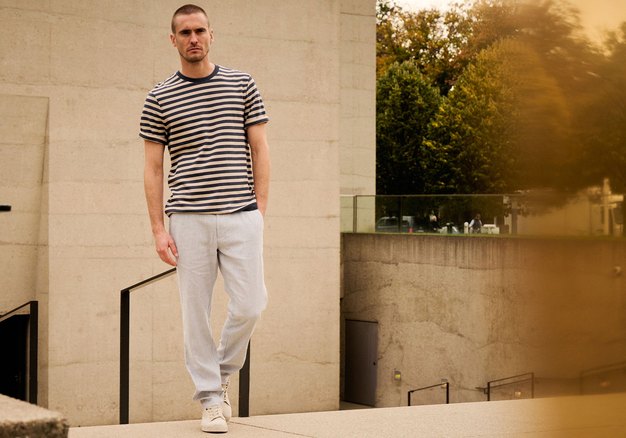 A man wearing a striped T-shirt and light linen pants walking outdoors in a modern urban setting.