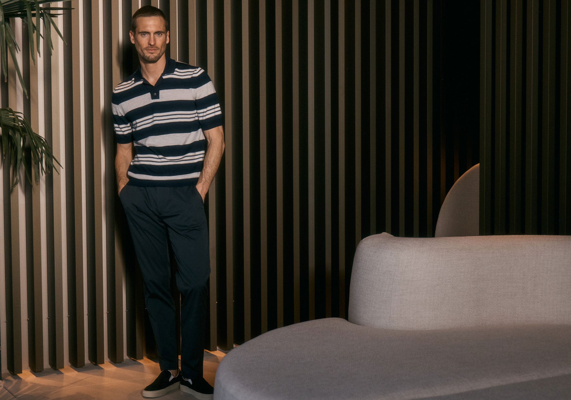 A man in a striped polo shirt and dark trousers stands casually against a modern slatted wall.