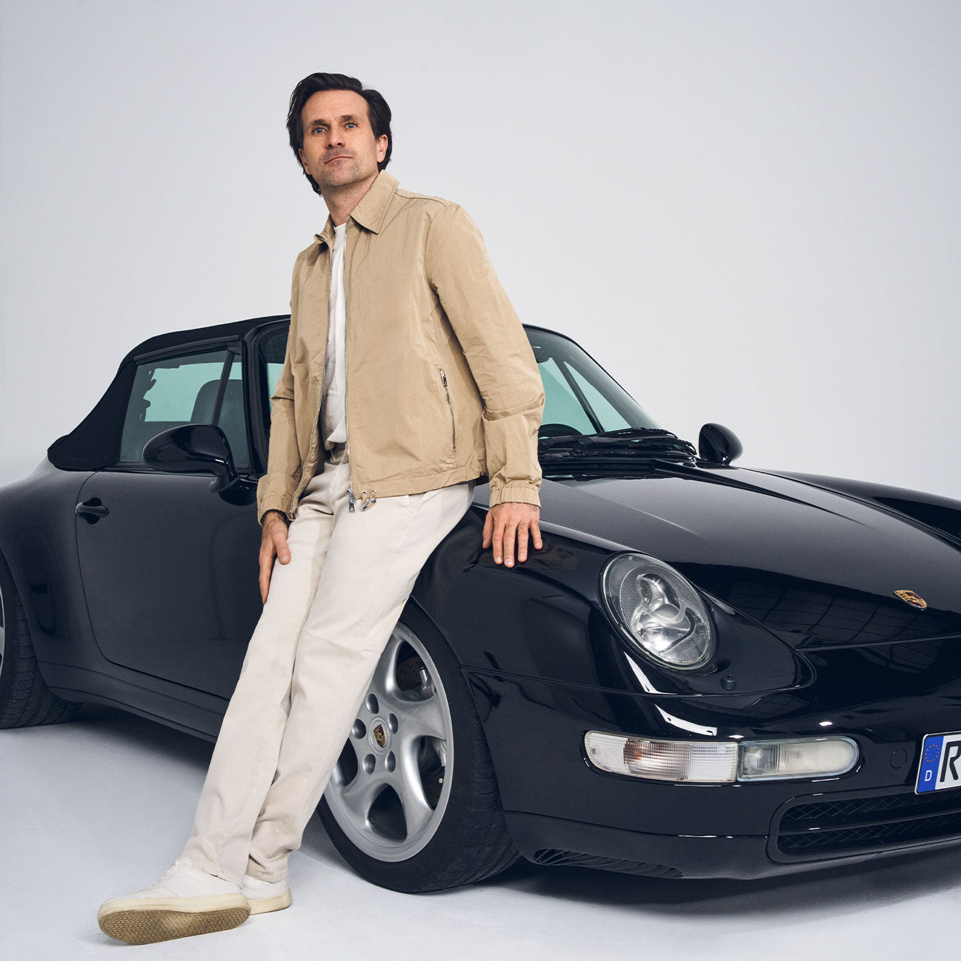 Man wearing a beige jacket and light-colored pants leans casually against a black sports car in a bright studio, looking confidently toward the camera.