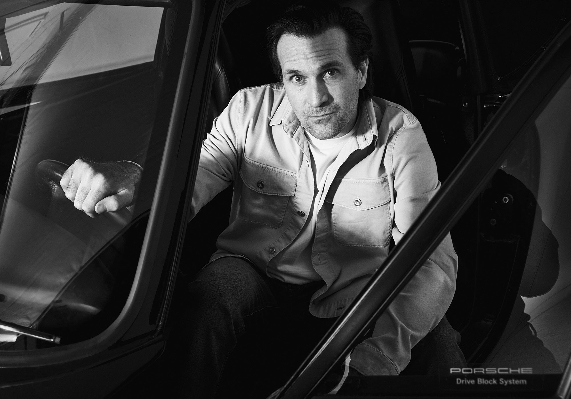 Black-and-white portrait of a man sitting inside a car, leaning slightly forward and looking directly at the camera.
