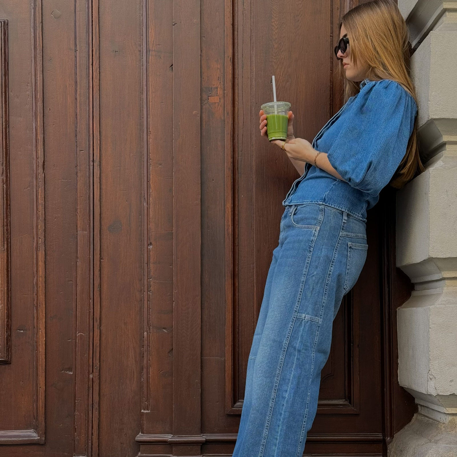 A woman in a blue denim outfit leans casually against a wooden door, holding a green drink in a plastic cup. She wears sunglasses and stands in a relaxed pose, highlighting the fit of her jeans.