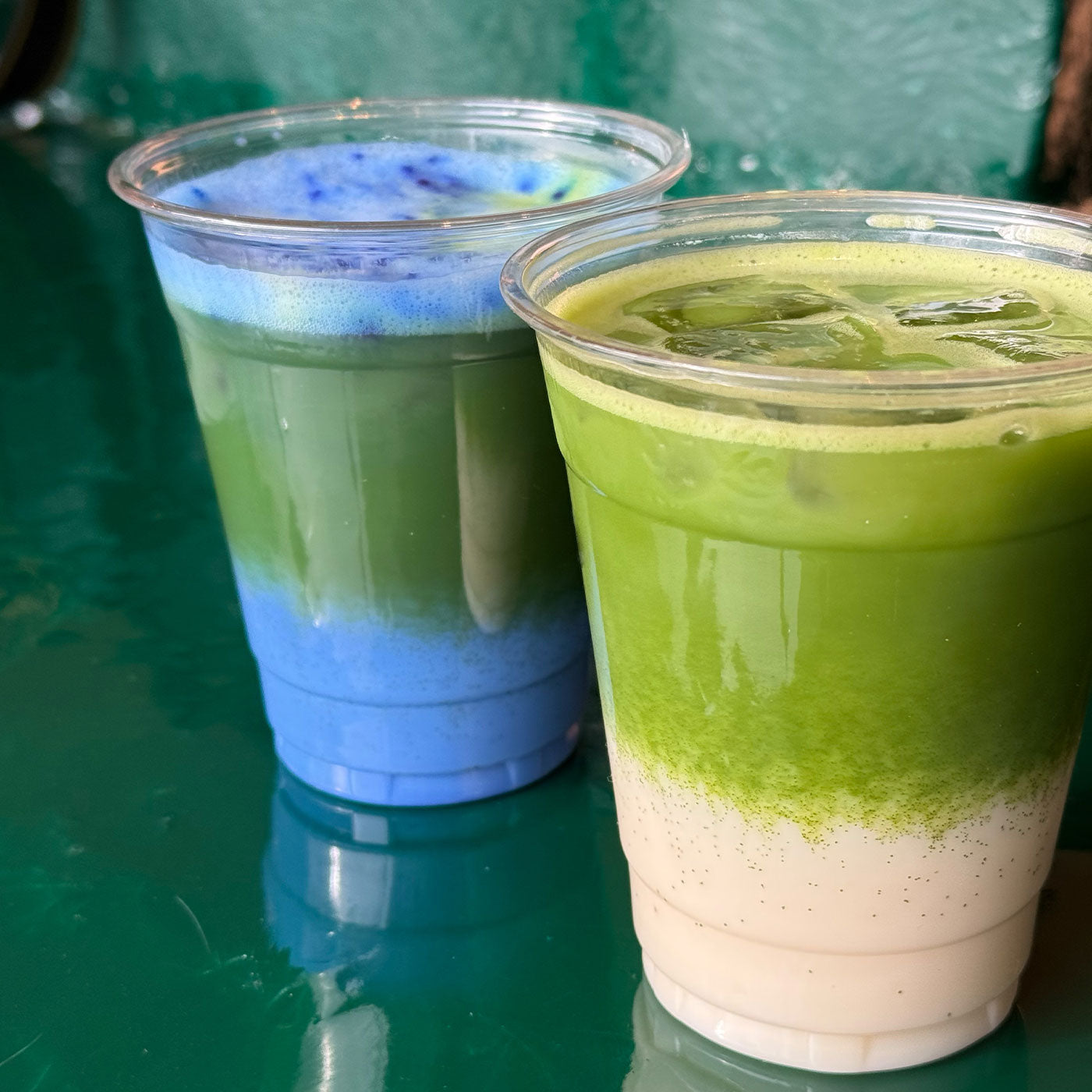 Two iced matcha drinks in clear cups on a green table, layered with milk and matcha foam.