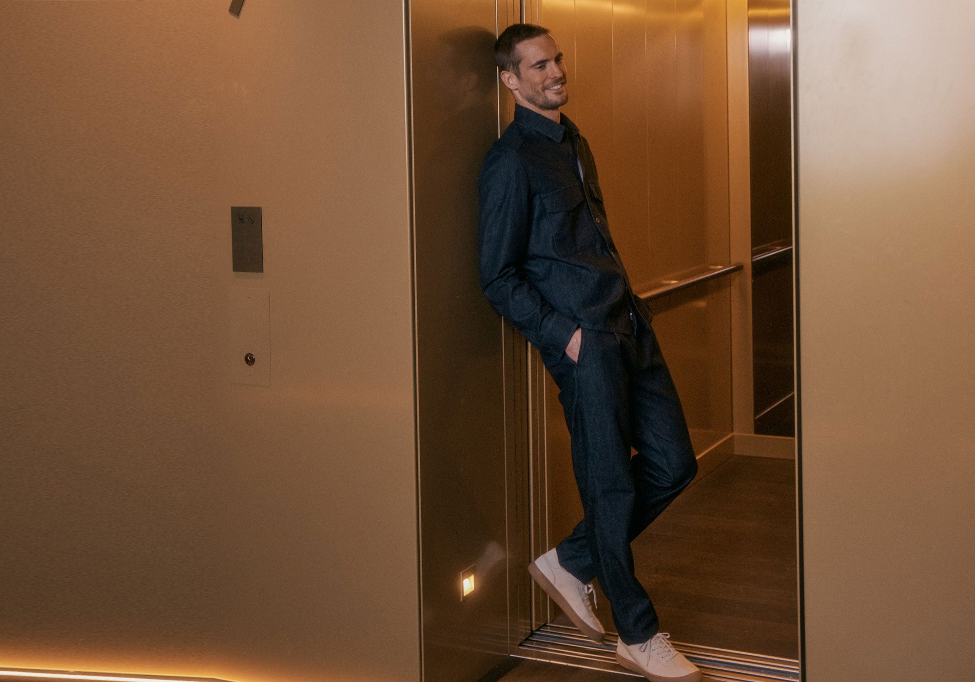 A man in a dark denim outfit leans casually in an elevator, wearing white sneakers.