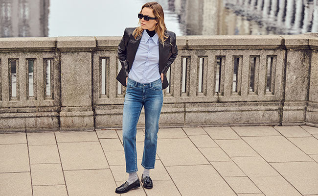 Woman in sunglasses wearing a dark blazer, light blue shirt, blue jeans Carrie Straight and loafers, standing by a stone railing beside water.